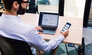 mężczyzna siedzący przy biurku aplikacja transakcyjna na telefonie i laptopie